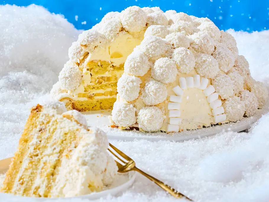 Kuppelförmiger Kokos-Iglu-Maulwurfkuchen, mit Kokoskugeln dekoriert und cremiger Füllung, auf einer weißen Platte angerichtet, daneben ein angeschnittenes Kuchenstück auf einem runden Teller mit goldener Gabel.