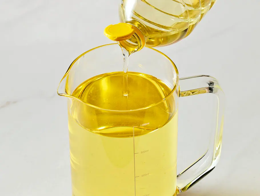 Klare Glas-Messkanne, gefüllt mit goldgelbem Pflanzenöl, in das gerade weiteres Öl aus einer Flasche gegossen wird. Das Gefäß steht auf einer hellen, glatten Unterlage.