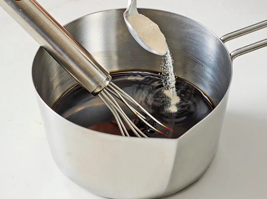 In einen kleinen Edelstahltopf mit dunklem Balsamico werden Granulat oder Pulver von einem Löffel hinzugegeben. Ein Schneebesen liegt bereit im Topf, der auf einer glatten, hellen Unterlage steht.