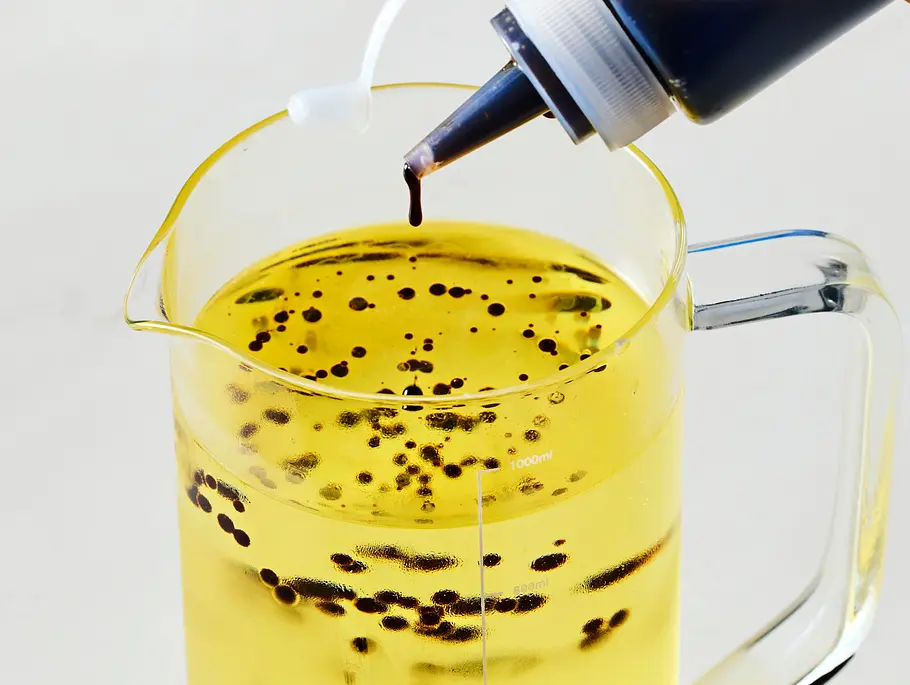 Dunkle Tropfen einer Balsamico-Mischung werden aus einer Dosierflasche in eine große Glas-Messkanne mit klarem, kaltem Pflanzenöl geträufelt. Die Kanone steht auf einer hellen Arbeitsfläche.