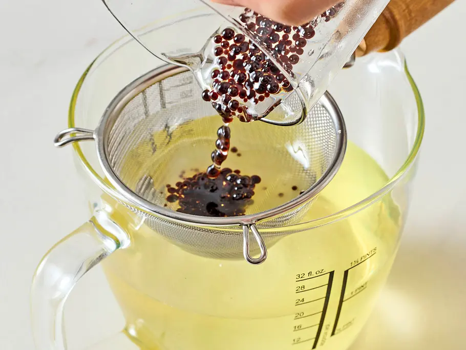 Dunkle Balsamico-Perlen werden aus einem Glas durch ein feines Sieb in eine große Glas-Messkanne gegossen, die mit hellem Öl gefüllt ist. Die Kanne steht auf einer hellen, glatten Arbeitsfläche.