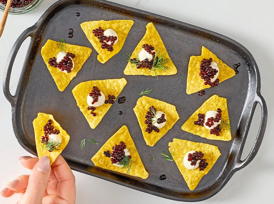 Knusprige, goldgelbe Dreiecke aus Maischips, jeweils mit einem Klecks Creme, kleinen schwarzen Balsamico-Perlen und etwas frischem Dill garniert, angerichtet auf einer dunklen Servierplatte. Daneben steht ein Glas mit den glänzenden Balsamico-Perlen und einem Holzlöffel. Eine Hand greift nach einem der belegten Chips.