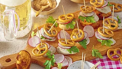 Weißwurst-Scheiben auf Holzbrett, belegt mit Radieschenscheiben, Petersilie und kleinen Brezeln, mit Zahnstochern fixiert. - Foto: LECKER @ Bauer Media Group