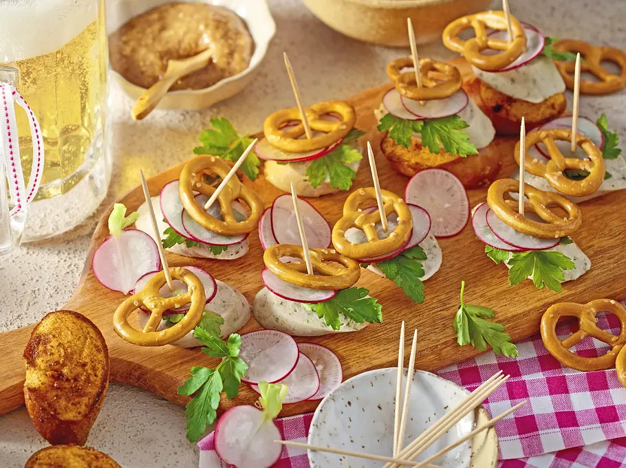 Weißwurst-Scheiben auf Holzbrett, belegt mit Radieschenscheiben, Petersilie und kleinen Brezeln, mit Zahnstochern fixiert.