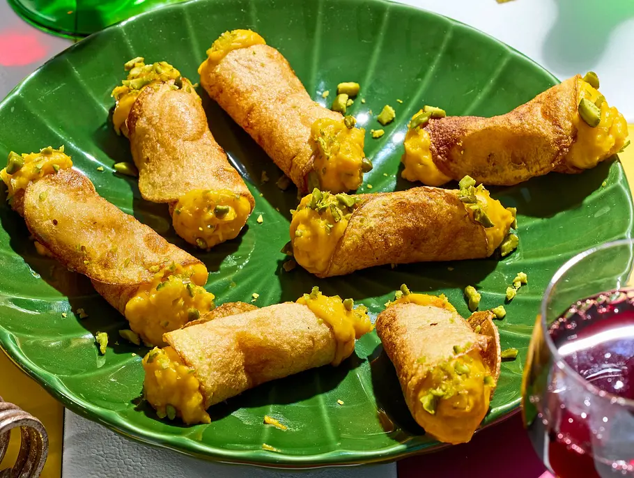 Acht herzhafte Cannoli mit goldgelber Füllung auf einem grünen, blattförmigen Teller, dazu stilvolle Weingläser mit Rotwein auf einem bunt karierten Tisch.