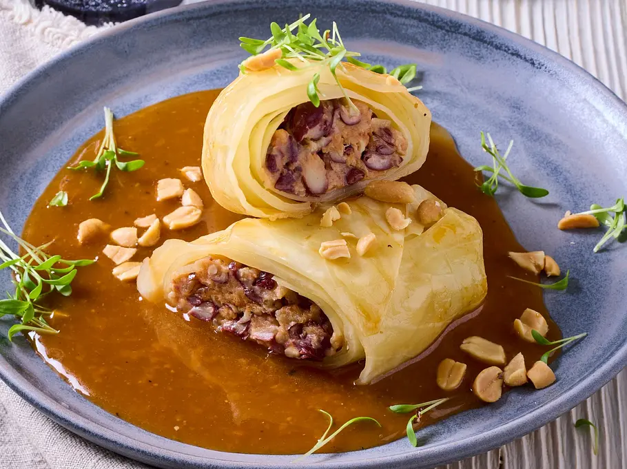 Zarte vegane Kohlrouladen, gefüllt mit einer herzhaften Bohnenmischung, in einer aromatischen braunen Soße, serviert auf einem grauen Teller und garniert mit frischen Sprossen und gehackten Erdnüssen.