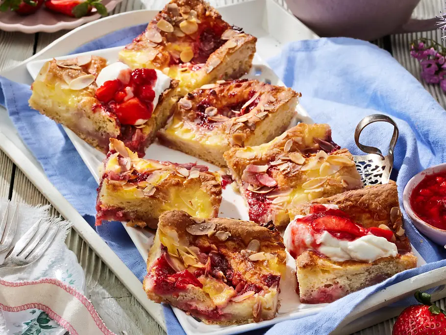 Mehrere Stücke Erdbeer-Butterkuchen mit goldbrauner Kruste, cremiger Puddingfüllung, Erdbeerstückchen und gerösteten Mandelblättchen, angerichtet auf einer rechteckigen weißen Platte mit blauem Tuch.