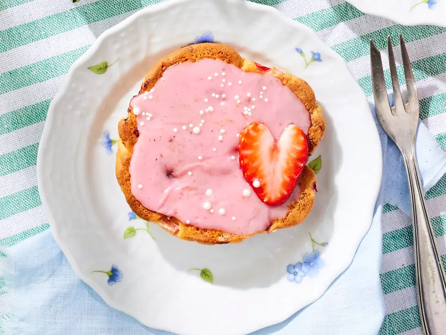 Drei Erdbeer-Amerikaner mit rosa Erdbeer-Zuckerguss, einzelnen Zuckerperlen und frischen Erdbeerscheiben, serviert auf kleinen weißen Tellern mit Blumenmotiv.