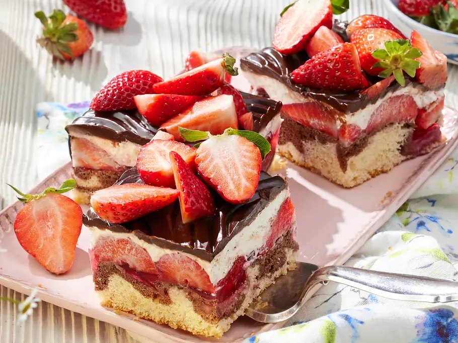 Zwei Stück Erdbeer-Donauwelle mit heller und dunkler Biskuit-Schicht, cremiger Füllung, Erdbeeren und Schokoguss, dekoriert mit frischen Erdbeeren auf einer rosa Platte.