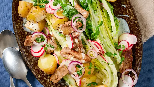 Kartoffelsalat mit halbierten kleinen Kartoffeln, frischem Römersalat, Rinderstreifen, Joghurtdressing und Radieschenscheiben auf einem ovalen, dunkel gemusterten Teller. Daneben liegt eine beigefarbene Serviette mit Besteck. - Foto: StockFood Studios / Meike Bergmann / House of Food