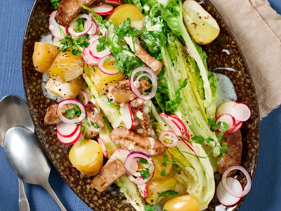 Kartoffelsalat mit Joghurtdressing und Rinderstreifen Kartoffelsalat mit halbierten kleinen Kartoffeln, frischem Römersalat, Rinderstreifen, Joghurtdressing und Radieschenscheiben auf einem ovalen, dunkel gemusterten Teller. Daneben liegt eine beigefarbene Serviette mit Besteck.