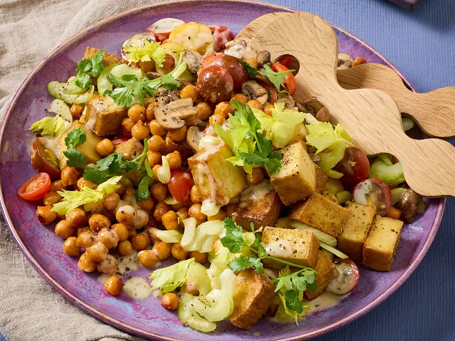 Lauwarmer Kichererbsen-Tofu-Salat Rezept Lauwarmer Salat aus gebratenem Tofu, gerösteten Kichererbsen, Sellerie, Champignons und Kirschtomaten auf einem lila Teller, garniert mit frischen Kräutern. Daneben Holzsalatbesteck auf einer Leinenserviette.