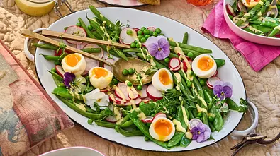 Frischer Gartensalat mit halbierten Eiern, Radieschenscheiben, grünen Bohnen, Erbsen, Spargel und essbaren Blüten auf einer weißen Platte, garniert mit einem cremigen Dressing. - Foto: LECKER @ Bauer Media Group