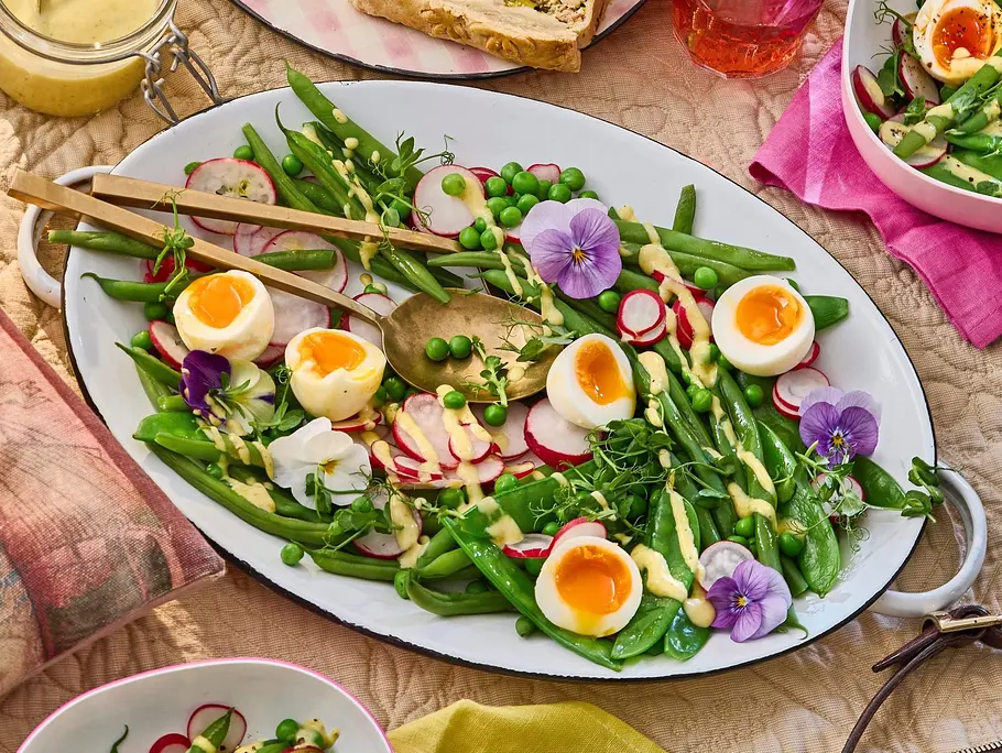 Frischer Gartensalat mit halbierten Eiern, Radieschenscheiben, grünen Bohnen, Erbsen, Spargel und essbaren Blüten auf einer weißen Platte, garniert mit einem cremigen Dressing.