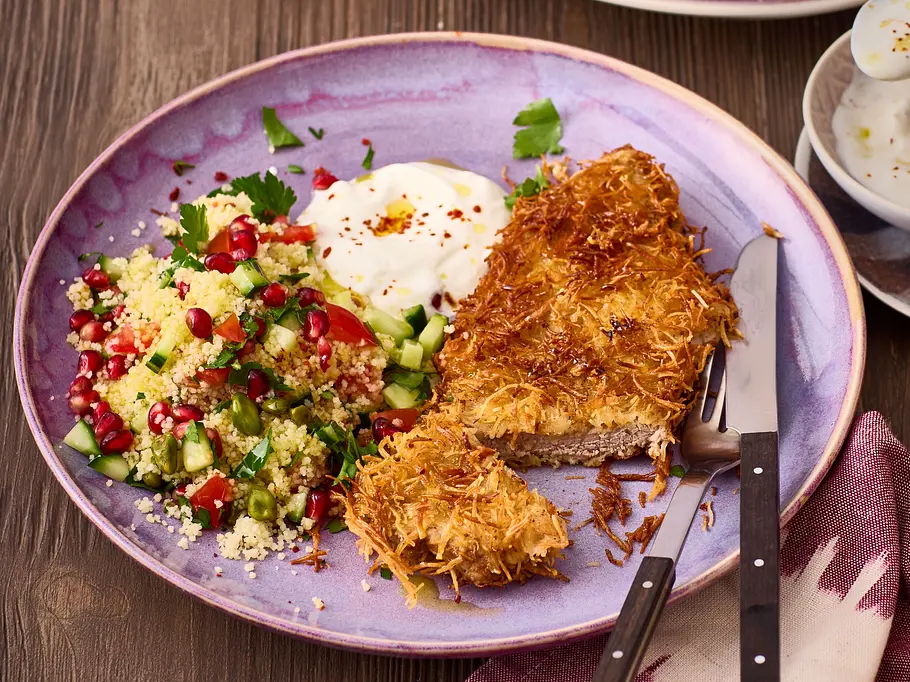 Knuspriges, in Kadayif-Teig paniertes Schnitzel auf einem violetten Teller, dazu bunter Couscous-Salat mit Granatapfelkernen und ein Klecks cremiger Joghurt-Dip.
