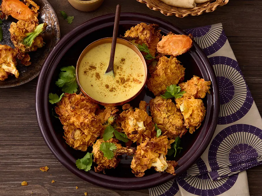 Knuspergemüse im Cornflakes-Mantel Knusprig frittiertes Gemüse im goldenen Cornflakes-Mantel auf einem dunklen Teller, serviert mit cremigem Dip in einer Schale und frischem Fladenbrot im Korb.
