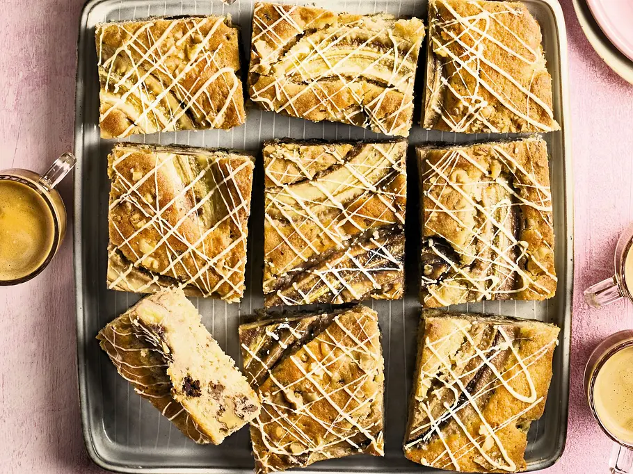 Neun saftige, hellbraune Bananenblondies mit sichtbaren Schokostücken und feinem weißen Schokoladenguss liegen in einem Backblech auf einem rosa Tisch. Rundherum stehen vier kleine Tassen mit Espresso sowie ein Stapel rosa Pastellteller.