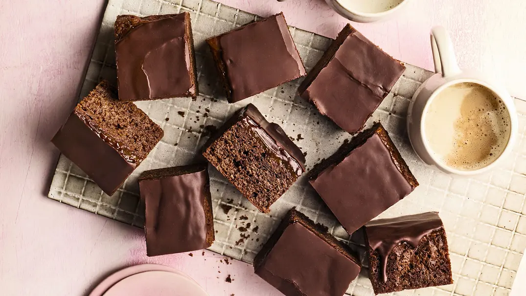 Mehrere saftige Sacher-Brownies mit glänzendem Schokoladenguss liegen auf einem weißen Gitter auf rosa Untergrund. Daneben stehen zwei Tassen cremiger Kaffee und ein Stapel rosafarbener Teller. - Foto: LECKER @ Bauer Media Group Mehrere saftige Sacher-Brownies mit glänzendem Schokoladenguss liegen auf einem weißen Gitter auf rosa Untergrund. Daneben stehen zwei Tassen cremiger Kaffee und ein Stapel rosafarbener Teller. - Foto: LECKER @ Bauer Media Group