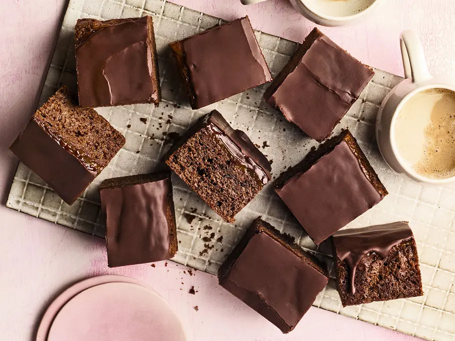 Mehrere saftige Sacher-Brownies mit glänzendem Schokoladenguss liegen auf einem weißen Gitter auf rosa Untergrund. Daneben stehen zwei Tassen cremiger Kaffee und ein Stapel rosafarbener Teller.