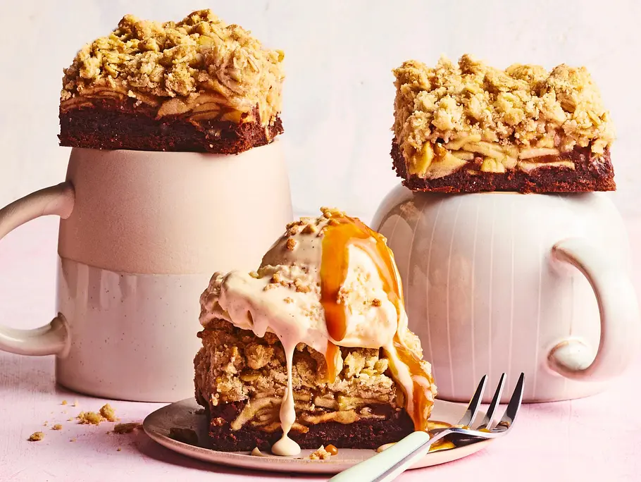 Zwei dicke Stücke Apfel-Strownies mit goldbraunem Knusper-Crumble auf Kaffeetassen angerichtet, davor ein weiteres Stück auf einem Teller, getoppt mit einer Kugel Eis und karamelliger Sauce, daneben eine kleine Gabel auf rosa Untergrund.