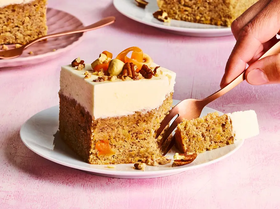 Drei saftige Carrot Cake Blondies mit heller Cremehaube, dekoriert mit knackigen Nüssen und Möhrenraspeln, serviert auf weißen und rosa Tellern. Im Hintergrund stehen zwei Gläser mit cremigem Kaffee, auf einem zartrosa Tisch.