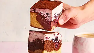 Drei große, üppige Stücke Donauwelle Tricolore mit hellgelbem Biskuitboden, dunkler Schokoladenschicht, beerig-fruchtiger Creme und Kakao bestäubt, gestapelt auf einem rechteckigen Brett. Daneben steht eine Tasse Cappuccino auf einem zartrosa Untergrund. - Foto: LECKER @ Bauer Media Group