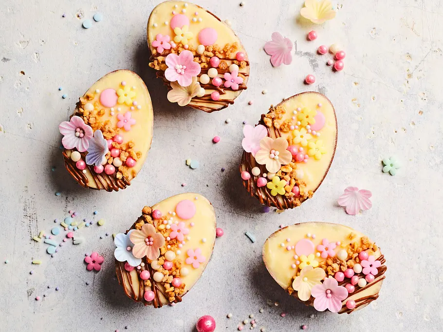 Fünf halbe Schoko-Vanillecreme-Eier mit zartem Schokoladenrand und heller Glasur, dekoriert mit rosa und gelben Zuckerblüten, kleinen Perlen und knusprigen Streuseln, arrangiert auf einer hellen, leicht strukturierten Unterlage.