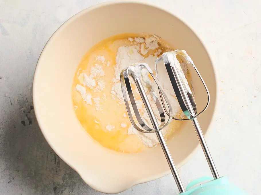 Vanillecremepulver wird in einer hellen Rührschüssel mit Milch und Eierlikör vermischt, daneben liegt ein Handmixer mit eingesetzten Rührbesen auf einer hellen Arbeitsfläche.
