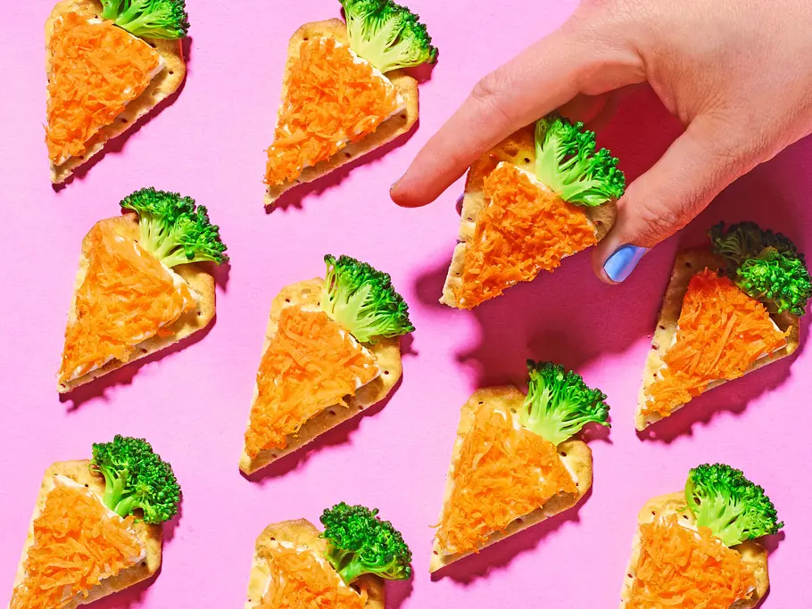 Dreieckige Cracker sind mit Frischkäse bestrichen und mit geraspelter Möhre belegt, sodass sie wie kleine Karotten aussehen; oben steckt jeweils ein Brokkoliröschen als „Karottengrün“. Eine Hand greift nach einem der Cracker vor pinkfarbenem Hintergrund.