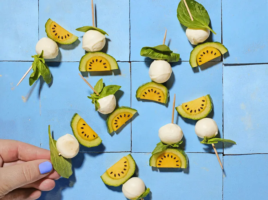 „Melonen“-Caprese Mehrere kleine Spieße mit Minimozzarella, grün-gelber Tomate und frischem Basilikum auf blauen Keramikfliesen angerichtet.