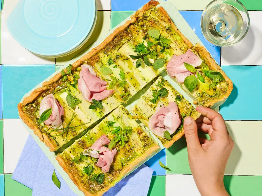 Eine rechteckige Spargel‑Kräuter‑Tarte mit Schinken wird serviert, während gerade ein Stück herausgenommen wird.