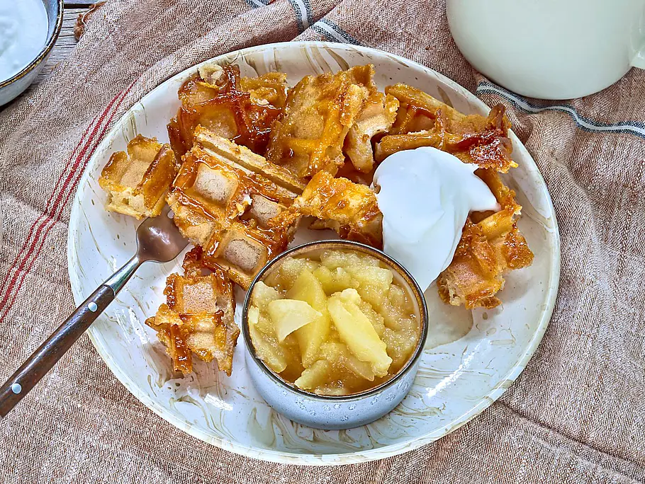 Knusprige Waffle Bites Knusprige Waffelschmarrn-Bites auf zwei Tellern, serviert mit Klecksen Sahne und Schälchen Apfelmus und Blaubeeren, auf einem Holztisch mit beigem Leinentuch, dazu zwei Tassen Kaffee.