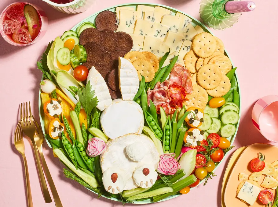 Buntes Snackboard für den Osterbrunch in Hasenform angerichtet, mit Käse, Crackern, Salami, frischem Gemüse wie Gurken, Tomaten und Erbsenschoten sowie Dip und kleinen Blüten, serviert auf einer großen runden Platte.