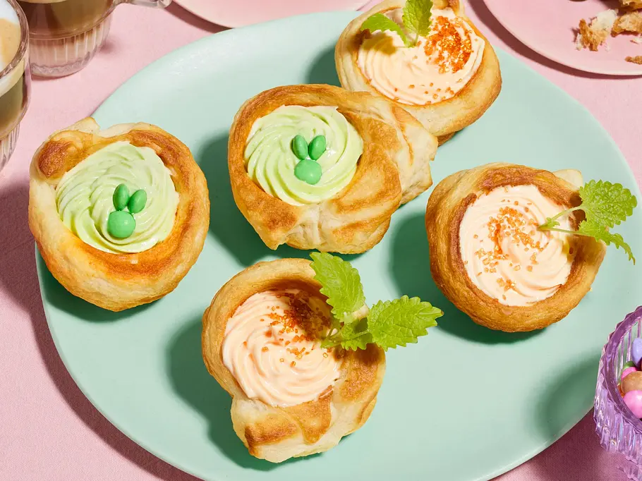 Goldbraun gebackene Croissant‑Nester, gefüllt mit farbiger Creme in Pastelltönen wie Grün und Apricot, dekoriert mit kleinen Zuckerstreuseln und frischer Minze, angerichtet auf einem Teller.