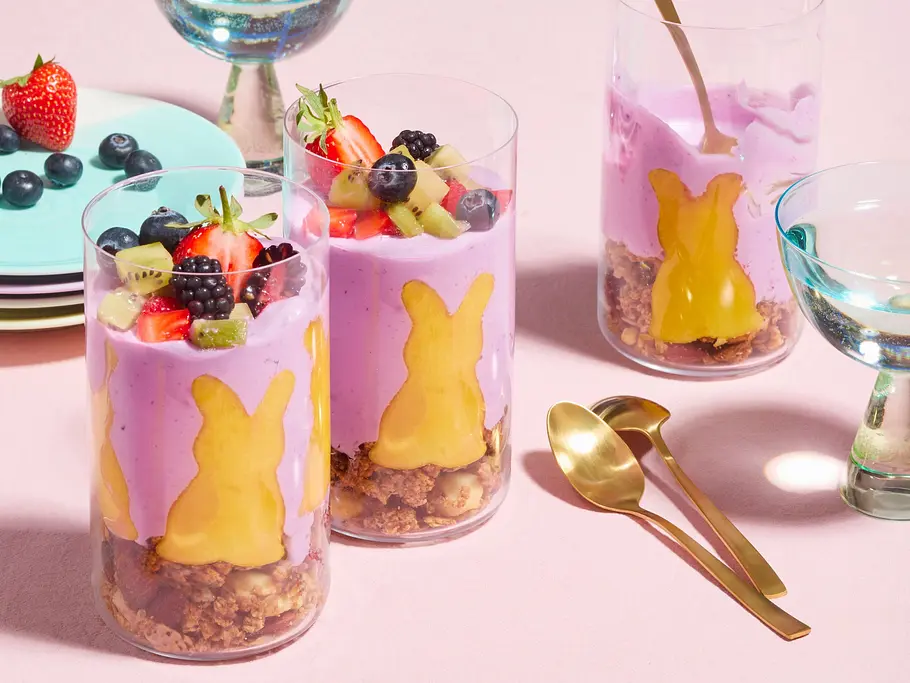 Frühstücks-Müsli im Glas mit knusprigem Granola, cremigem Joghurt und frischen Beeren, verziert mit einem Mango-Bunny in Hasenform, serviert in klaren Gläsern mit goldenen Löffeln.