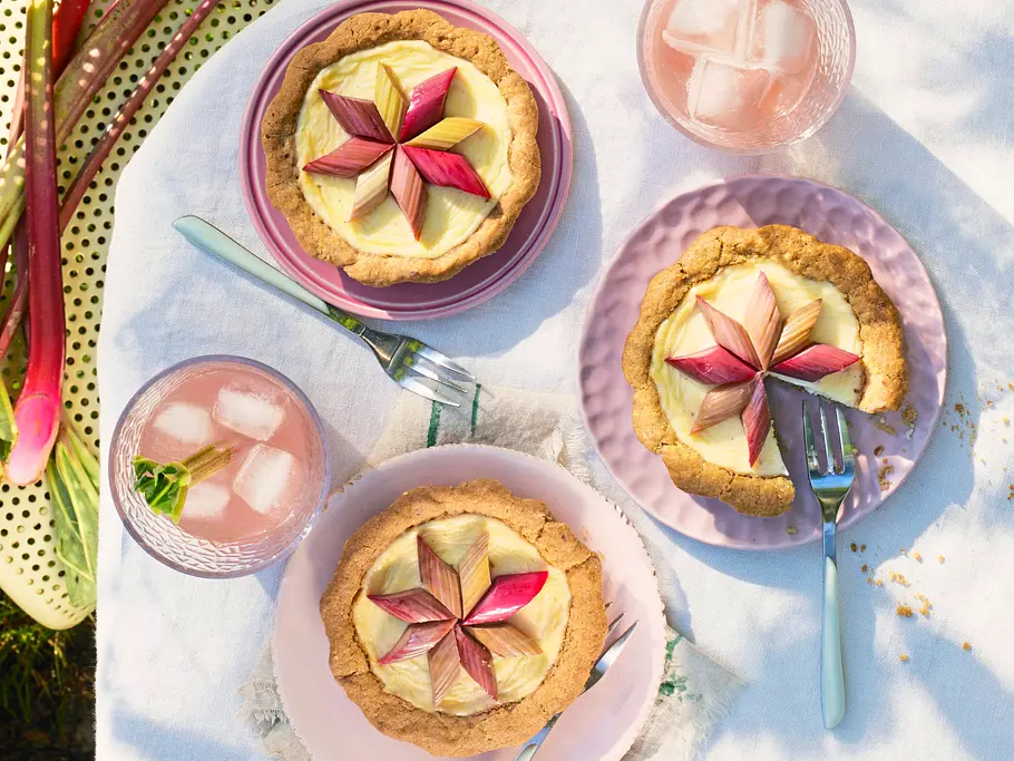 Drei kleine Mascarpone-Tartelettes mit goldbraunem Mürbeteig und cremiger Füllung, jeweils mit einem dekorativen Stern aus rosa und grünem Rhabarber belegt. Serviert auf zarten Pastelltellern mit Gabeln, daneben Gläser mit rosafarbenem Getränk und frische Rhabarberstangen auf einer hellen Unterlage im Freien.