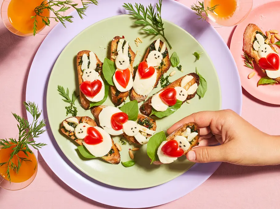 Geröstete Crostini mit Pesto, Mozzarella und Tomaten, dekoriert als kleine Häschen mit Basilikumblättern, angerichtet auf einem runden Teller.