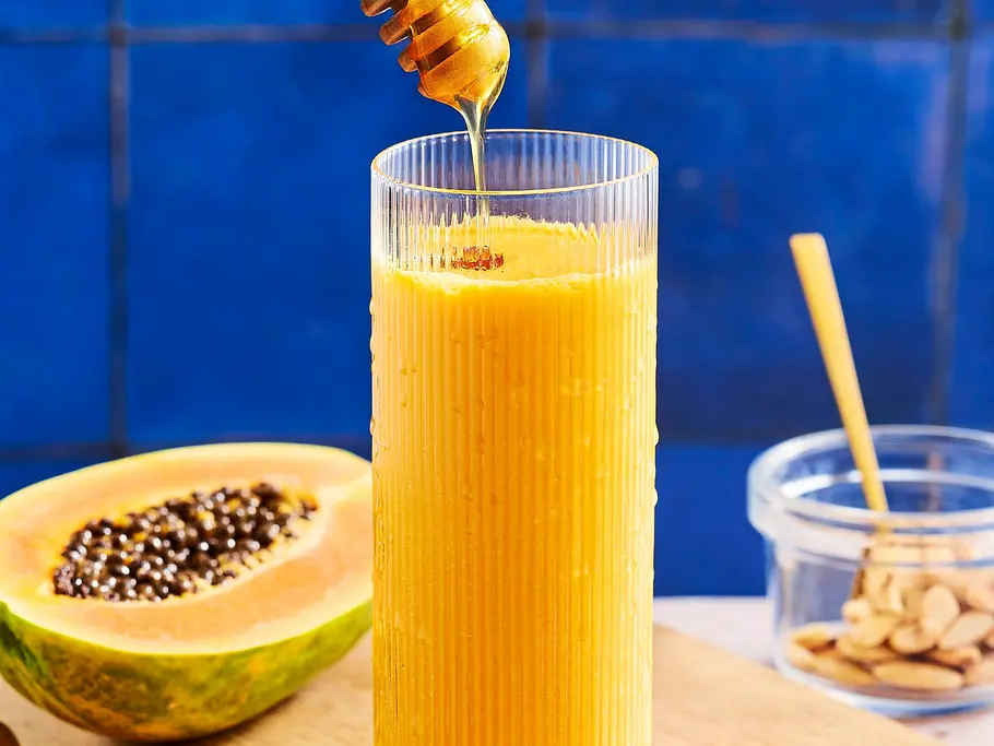 Sanddorn-Immun-Drink Gelber Sanddorn-Immun-Drink in einem hohen, geriffelten Glas auf einem Holzbrett, dekoriert mit frischem Honig, daneben Mandeln, Kurkumapulver und Orangenspalten.