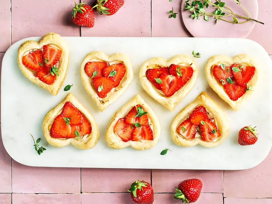 Mini-Ziegenkäse-Flammkuchen mit Erdbeeren Herzförmige Mini-Flammkuchen mit Ziegenkäse, frischen Erdbeeren und Thymian auf einer weißen Platte, dazu einige lose Erdbeeren und Thymianzweige.