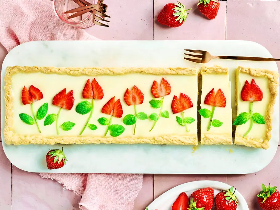 Rechteckige Pannacotta-Tarte auf einer weißen Platte, dekoriert mit Erdbeeren und Minzblättern in Blumenform, daneben frische Erdbeeren auf einem kleinen Teller.