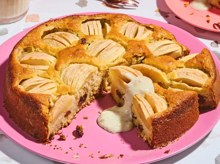 Runder, goldbrauner Apfelkuchen mit versunkenen Apfelspalten auf einem rosa Teller, daneben eine Stück auf einem weiteren rosa Teller mit Vanillesoße. Latte Macchiato im Glas und einige Kupfergabeln stehen daneben auf einer bunten, gemusterten Unterlage.
