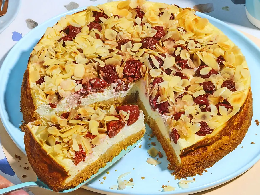Saftiger Kirsch-Quarkkuchen ohne Zucker auf einem hellblauen Teller, belegt mit Kirschen und gehobelten Mandeln, ein Stück ist angeschnitten und liegt auf einem kleinen Teller daneben.