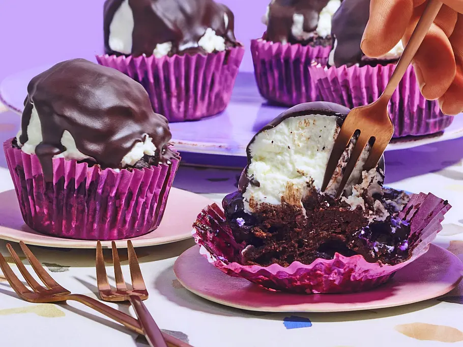 Schokokuss-Muffins ohne Zucker in lila Papierförmchen, umhüllt mit einer dicken Schokoladenschicht, auf kleinen Tellern angerichtet, ein Muffin wird mit einer Gabel angeschnitten.