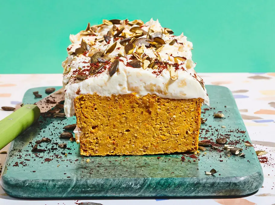 Saftiger Kürbis-Kastenkuchen mit heller Cremehaube, bestreut mit Kürbiskernen und dunklen Schokoraspeln, serviert auf einem grünen Brett.