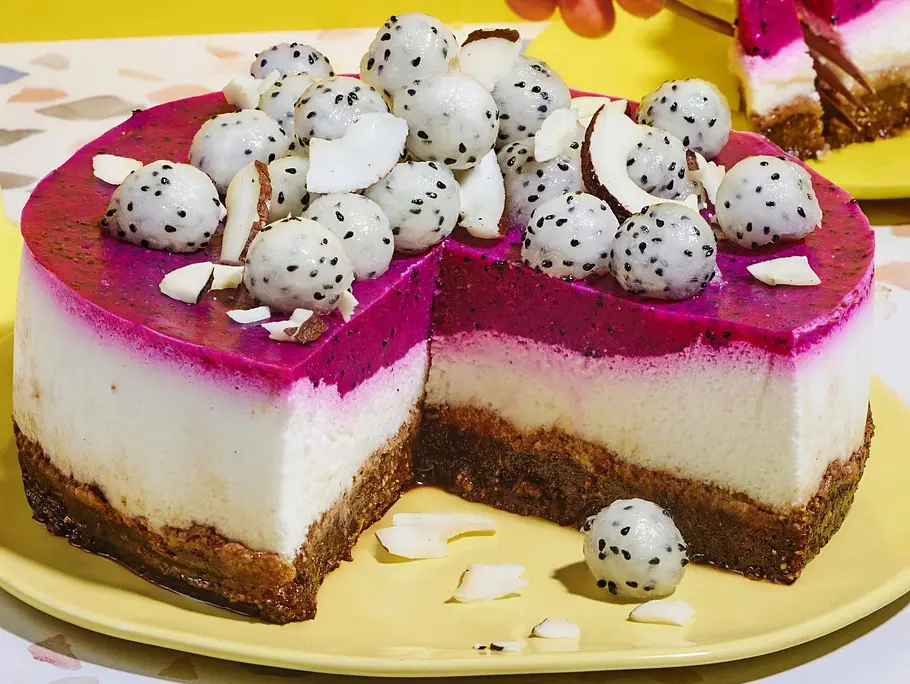 No-bake Torte mit leuchtender Pitahaya-Schicht, cremiger Kokosfüllung und knusprigem Boden, garniert mit frischer Drachenfrucht und Kokoschips, serviert auf gelbem Teller.