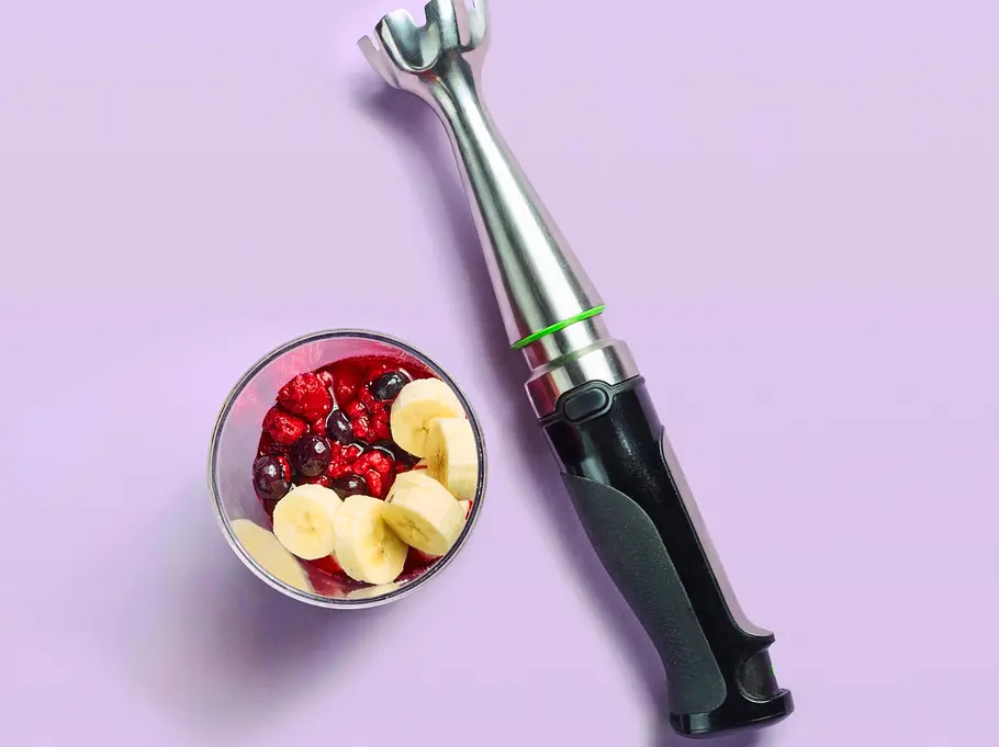 Ein Pürierstab liegt neben einem kleinen Glasbehälter, gefüllt mit frischen Bananenscheiben und roten Beeren.