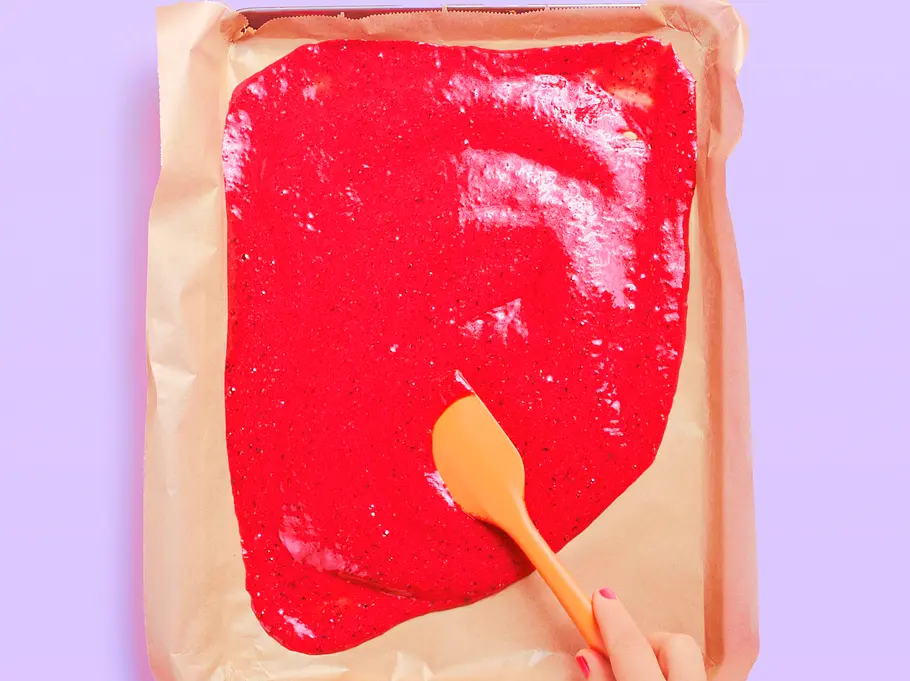 Ein Blech mit Backpapier, darauf eine gleichmäßig verstrichene, leuchtend rote Fruchtmasse; eine Hand verteilt die Masse mit einem orangefarbenen Spachtel.