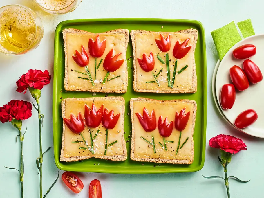 Vier Scheiben Käsetoast auf einem grünen Teller, belegt mit Tomatenstücken und Schnittlauch, die wie Tulpenblüten und Stängel angeordnet sind.