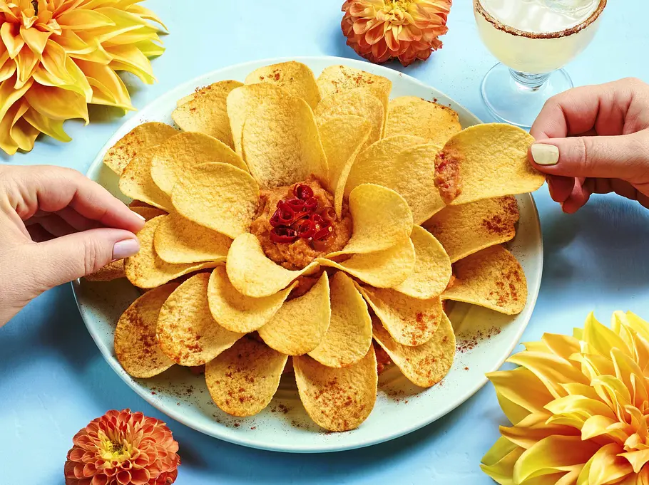 Kartoffelchips in Form einer Blüte auf einem weißen Teller, in der Mitte ein Dip mit Chili, serviert auf blauer Unterlage.