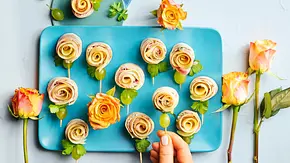 Mehrere Wrap-Röllchen in Rosenform auf einem türkisfarbenen Teller, dekoriert mit grünen Trauben und Petersilie, dazu gelbe Rosen als Dekoration. - Foto: LECKER @ Bauer Media Group